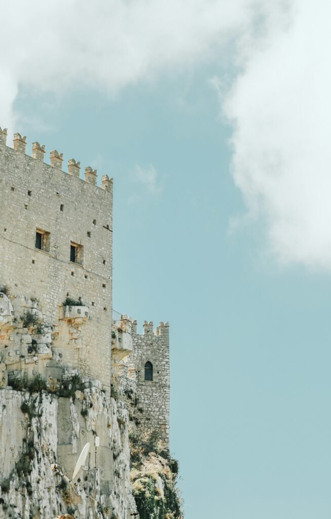 Caccamo Castle in Sicily, its pale stone walls and towers rising above the town under a clouded sky.