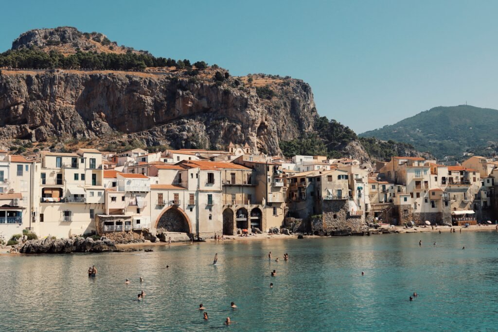 Swimmers in the sea below the seafront houses of Cefalù, with the rocky headland rising behind the town.