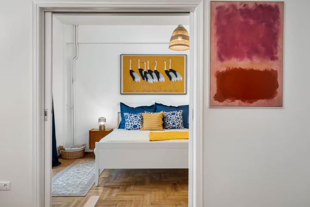 A bedroom seen through an open doorway, with blue and yellow cushions and large colour-field paintings on the walls.