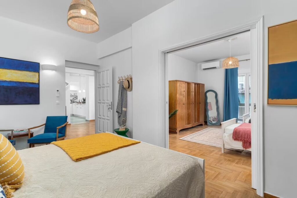 A double bedroom with a yellow throw across the bed and a blue abstract canvas, opening through to a second room.