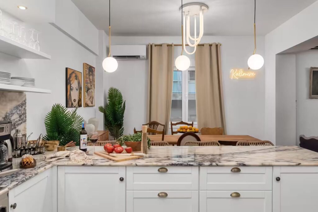 The kitchen island with herbs and fruit set out, and a small neon sign reading Kalimera on the far wall.