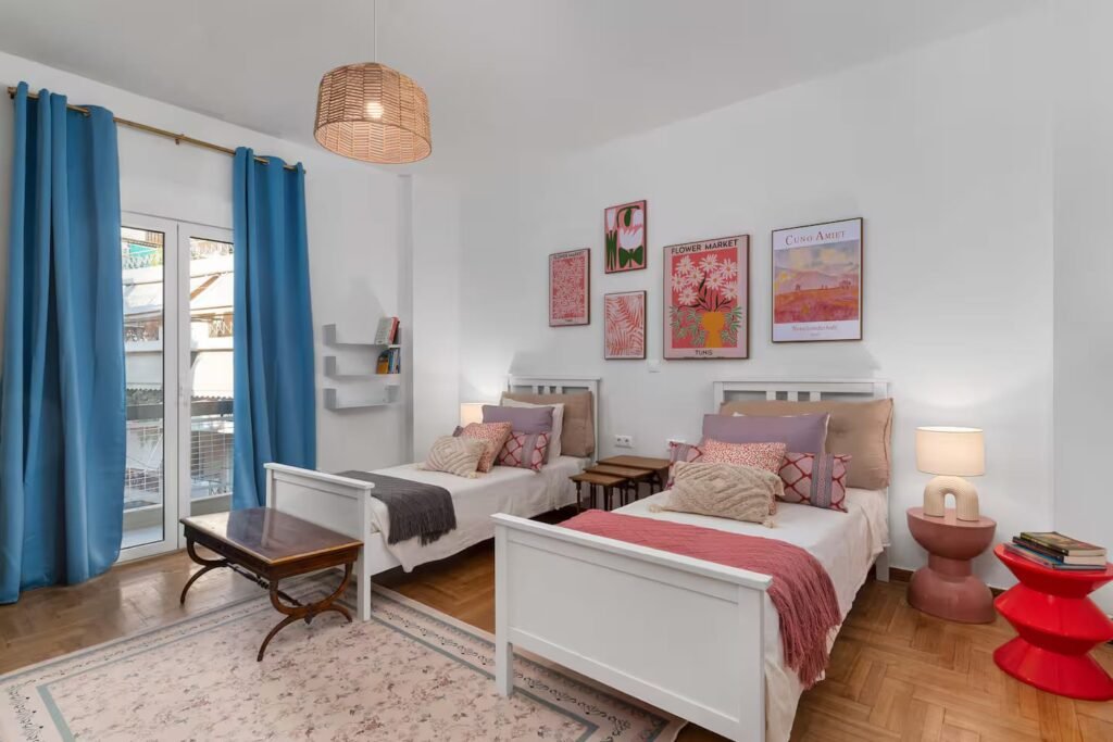 A twin bedroom with two single beds, blue curtains, a red side stool and framed prints above the beds.