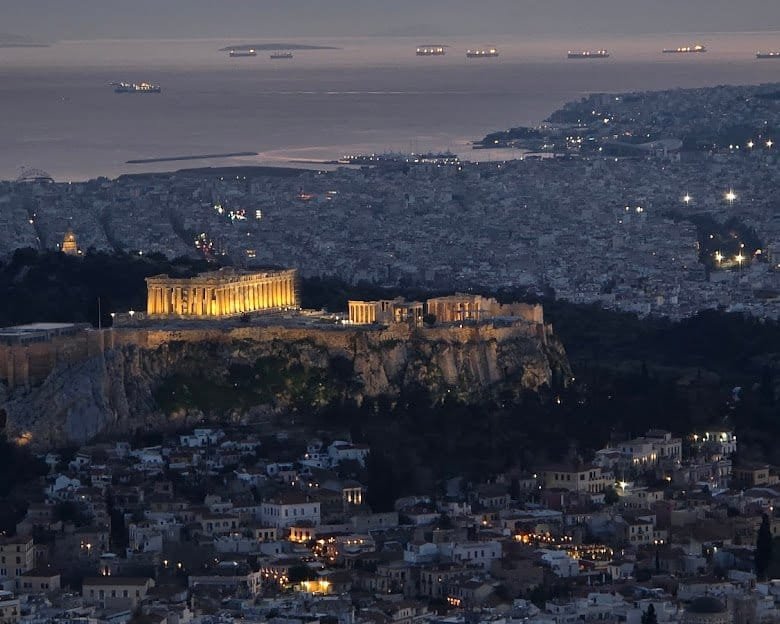 Athens at dusk from Lycabettus Hill, the floodlit Acropolis in the middle distance and the sea beyond.
