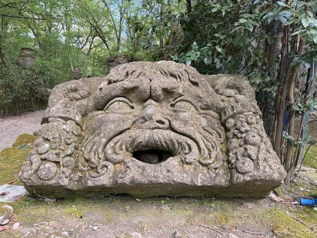 A vast carved stone face, its mouth wide open, half hidden by trees in the Sacro Bosco at Bomarzo.