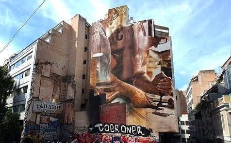A large mural covering the side wall of an Exarchia building above a graffitied shopfront.
