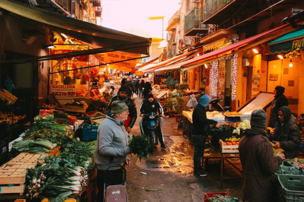 A busy Palermo street market at dusk, stalls piled with vegetables under lit awnings.