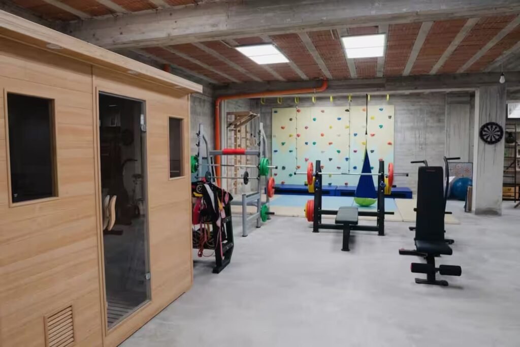 The basement gym: free weights and benches on rubber matting, a coloured climbing wall, and the sauna’s timber door.