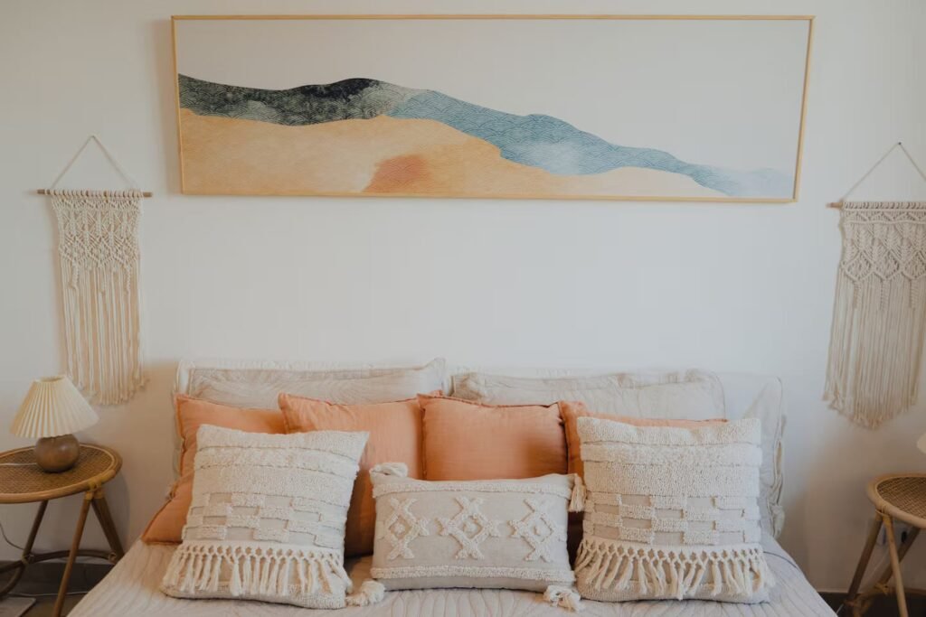 A bedroom dressed in cream and peach linens, with macramé wall hangings and a landscape painting above the bed.