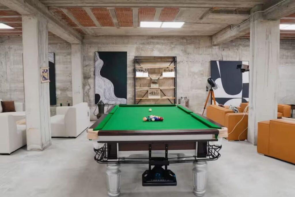 The games room, with a full-size pool table under pendant lights and armchairs against a concrete wall.