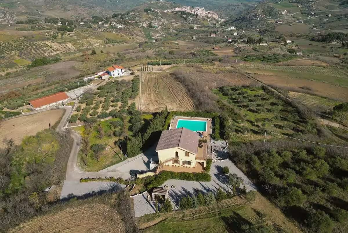 A wide aerial view showing the villa on its own hillside, with the surrounding Sicilian countryside falling away.