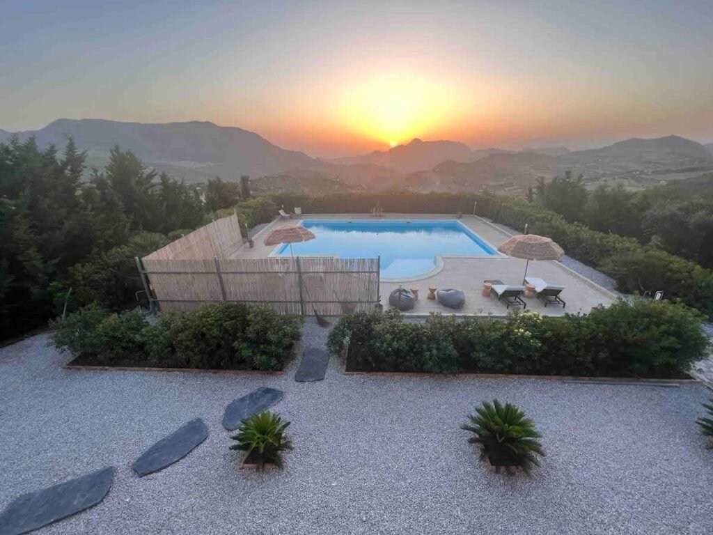 The swimming pool at dusk, its water still, with the sun setting behind the mountain ridge.