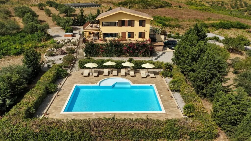 The villa from above: an ochre house on a terraced hillside beside a rectangular blue pool, ringed by olive trees.