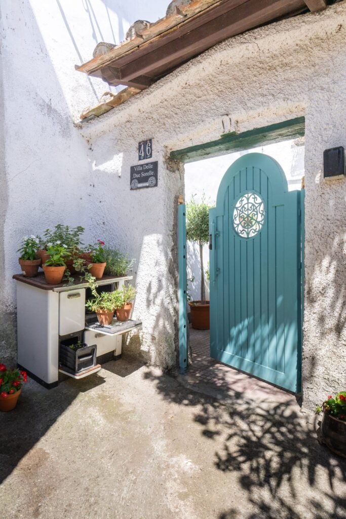 The villa’s teal-painted gate set into a whitewashed stone wall, with potted geraniums on the steps leading up to it.